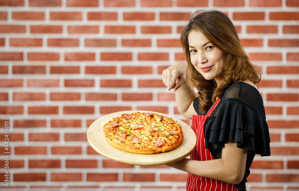 Asian beautiful woman wearing red apron, smiling and pointing finger for presenting homemade pizza with ham, bell peppers on wooden tray, standing in kitchen with background of brick wall with blank 