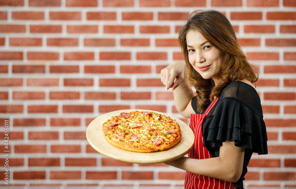 Asian beautiful woman wearing red apron, smiling and pointing finger for presenting homemade pizza with ham, bell peppers on wooden tray, standing in kitchen with background of brick wall with blank Asian beautiful woman wearing red apron, smiling and pointing finger for presenting homemade pizza with ham, bell peppers on wooden tray, standing in kitchen with background of brick wall with blank