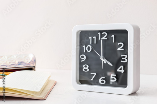 Stylish clock and notebooks on table near light wall
