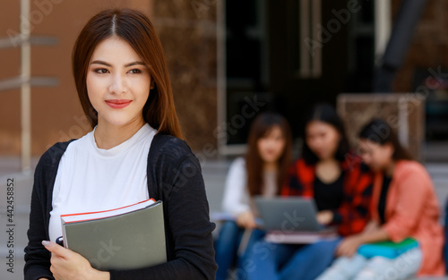 Young and beautiful Asian college student girls holding books, pose to camera with group of friends blur in background against school building. Learning and friendship of teens close friend concept