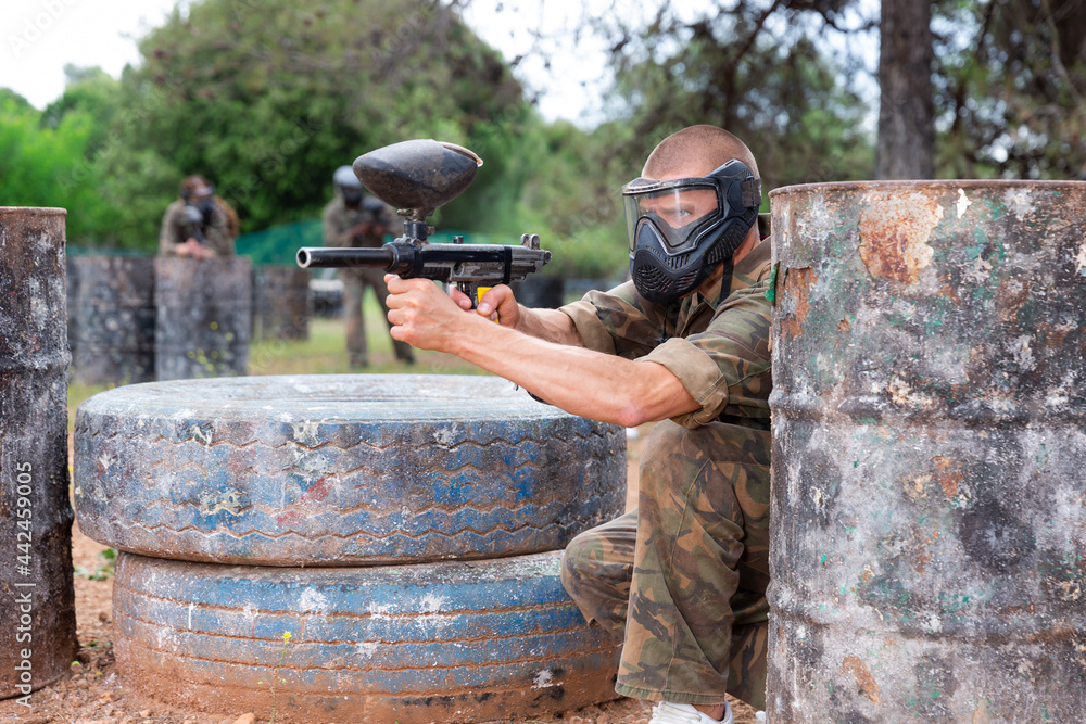 Fototapeta premium Male paintball player in protective mask standing with gun on paintball field