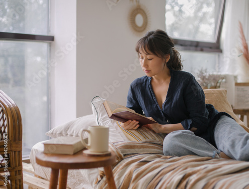 Young relaxed and happy Asian woman in casual outfit reading book and drinking tea while relaxing on sofa in cozy living room at home, enjoying free time on weekend at home