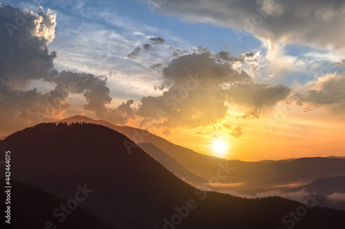 Sunset landscape of high mountain peaks and foggy valley under vibrant colorful evening sky. © bilanol