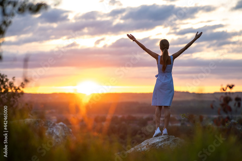 Dark silhouette of a young woman standing with raised up hands on a stone enjoying sunset view outdoors in summer.