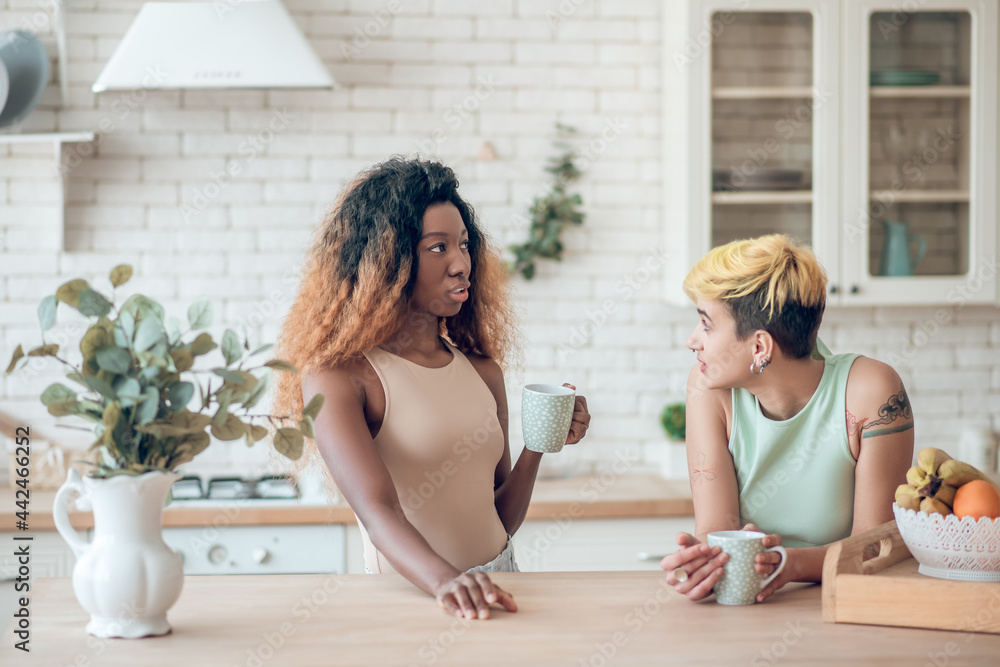 Two female friends chatting drinking coffee in kitchen Two female friends chatting drinking coffee in kitchen