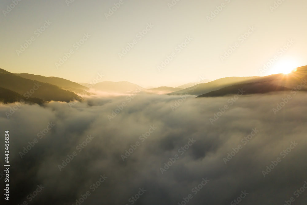 Aerial view of vibrant sunset over white dense foggy clouds with distant dark silhouettes of mountains on horizon.