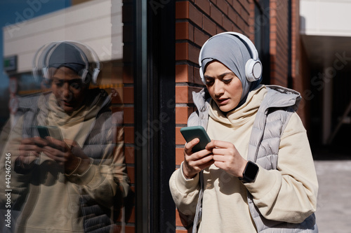 Waist up portrait of modern Middle-Eastern woman using smartphone in city lit by sunlight and wearing headphones, copy space