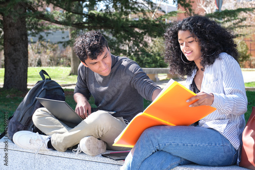 Two latin students studying from their lecture notes sitting on a wall outdoors. University life at campus.
