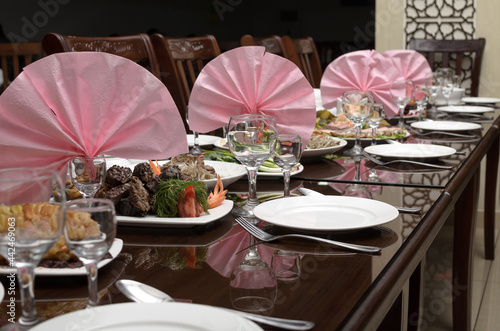 Wallpaper Mural Large brown festive banquet table is decorated with pink napkins and served with white plates, spoons, forks, wine glasses, festive salads and snacks from vegetables and meat are on the table Torontodigital.ca