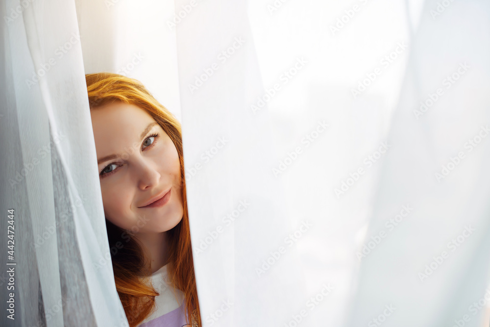 Cute young red-haired girl looks out from the white curtain, close-up, copy space. Portrait of beautiful smiling woman with perfect skin on a light blurry background. Cute young red-haired girl looks out from the white curtain, close-up, copy space. Portrait of beautiful smiling woman with perfect skin on a light blurry background.