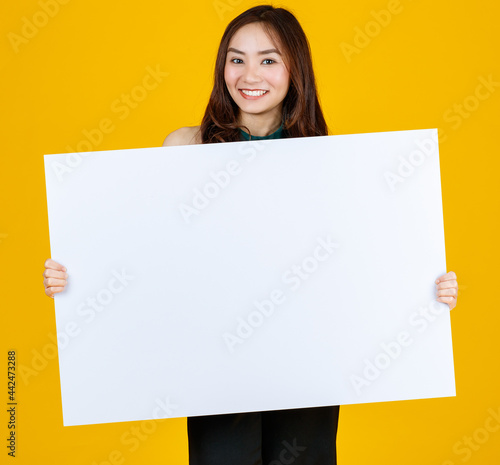 Cute and pretty curly hair Asian female brunette holding white blank board poses to camera with a joyful for advertising and banner use purpose, studio shot isolated on bright yellow background