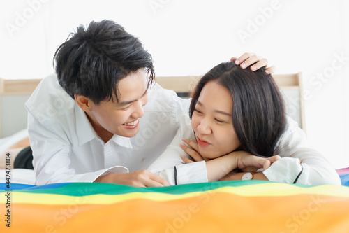 LGBTQ couple lovers, a handsome girl as a man or butch femme girl laying on a bed with the rainbow flag the symbol of LGBT, spending and sharing loving time in white bedroom with fun photo