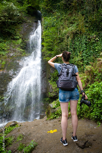 A slender brunette with a backpack and a camera stands near a picturesque waterfall.