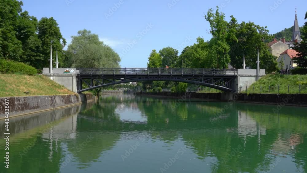 Beautiful city of Ljubljana, Slovenia in summer. Iron bridge over Ljubljanica river. Low angle view of calm river flowing in man made canal. Bright sunny day. Handheld, real time Beautiful city of Ljubljana, Slovenia in summer. Iron bridge over Ljubljanica river. Low angle view of calm river flowing in man made canal. Bright sunny day. Handheld, real time