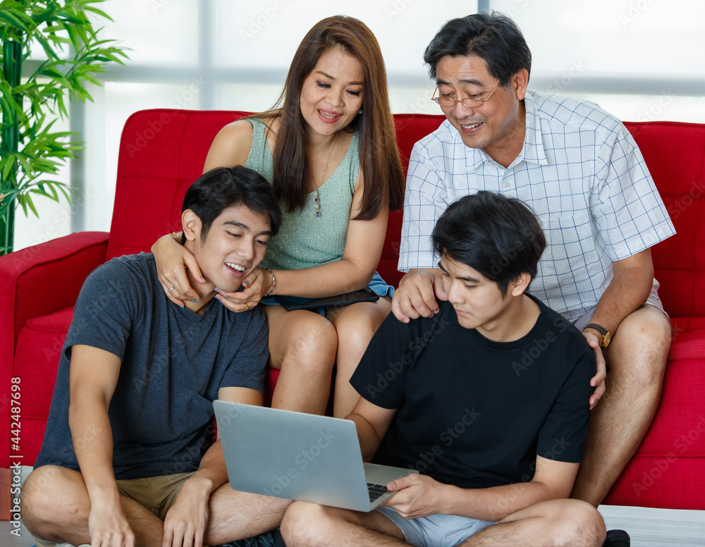 Fototapeta premium Four members Asian family consisting of parents, two adult sons happily spending time together during holidays, using laptop in comfortable living room at home. Presenting modern technology concept.