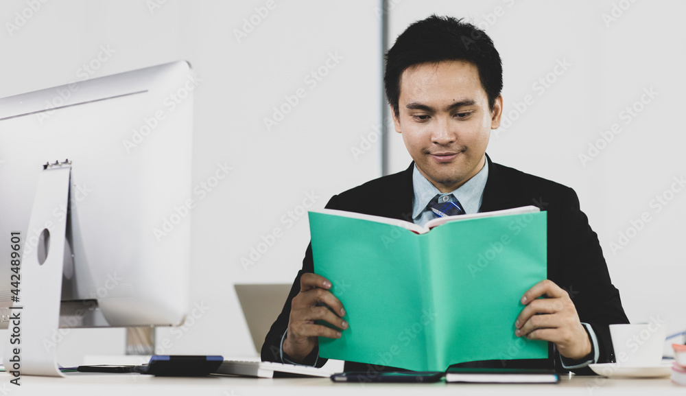 Portrait focus to adult asian businessman very smart and handsome hold and do reading note at work desk while feeling comfortable and happy smile on face. On desk have computer, calculator and tablet Portrait focus to adult asian businessman very smart and handsome hold and do reading note at work desk while feeling comfortable and happy smile on face. On desk have computer, calculator and tablet