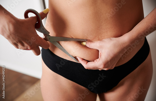 Close up of female hands cutting belly fat with scissors. Young woman in black underwear trying to get rid of excess weight. Concept of weight loss, diet and overweight.