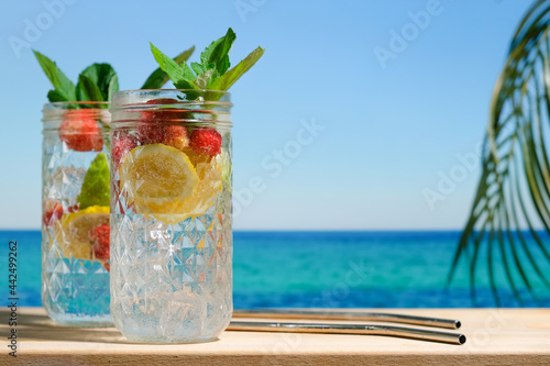 Summer cocktails, beach bar concept. Two drinking glasses of hard seltzer cocktail with strawberry, lemon and mint, beautiful sea on background. Summer resort concept.