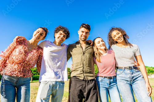 Multiracial group of college students embracing each other with arms around smiling looking at camera in a sunny day with blue sky at city park. Concept of equality and brotherhood among young people