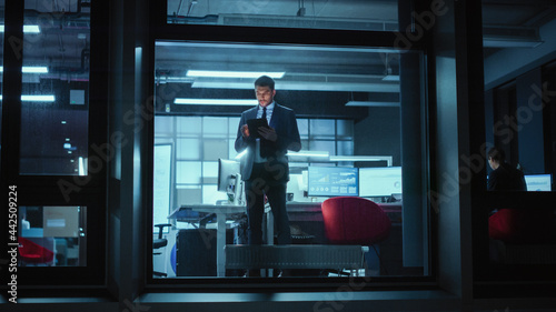 Shot from Outside: Businessman Working on a Digital Tablet Computer in the Office. Manager Checking Emails while Standing by the Window. Employee Planning Financial Tasks and Business Development. © Gorodenkoff