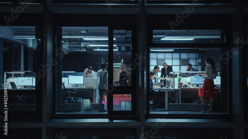 Shot from Outside the Window: Businessmen and Businesswomen Working in the Office. Managers and Specialists Doing Financial Business in the Evening. Employees Work on Computers and Delegate Tasks.
