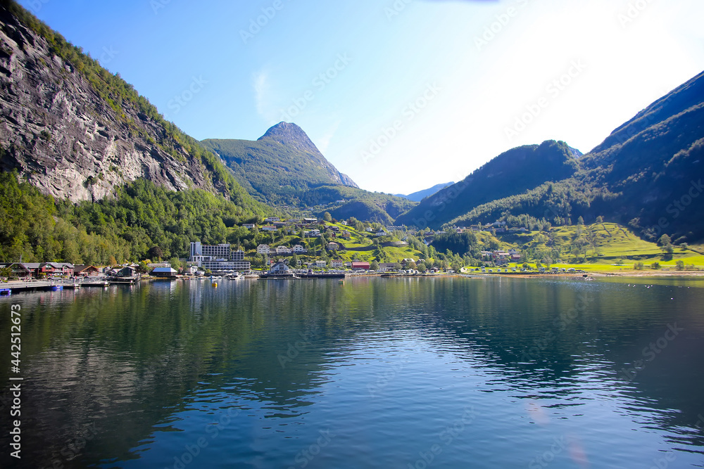 Beautiful view of Geiranger town from the water on a summer day. Reflections of the mountains in the fjord. lush green landscape with peaks towering over the village. Norway.
