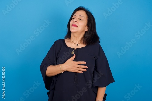 Satisfied smiling middle aged Arab woman standing against blue wall, keeps hands on belly, being in good mood after eating delicious supper, demonstrates she is full. Pleasant feeling in stomach.