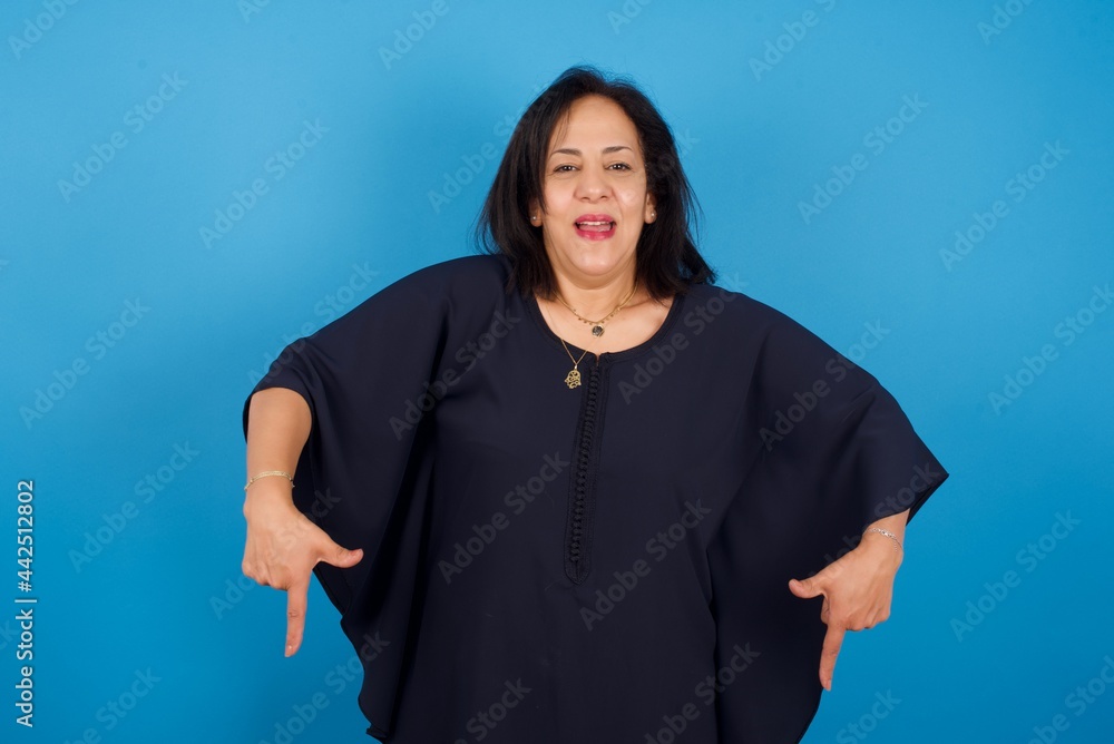 middle aged Arab woman standing against blue background with positive expression, points down with both index fingers, has broad interested smile. Look there, please. middle aged Arab woman standing against blue background with positive expression, points down with both index fingers, has broad interested smile. Look there, please.