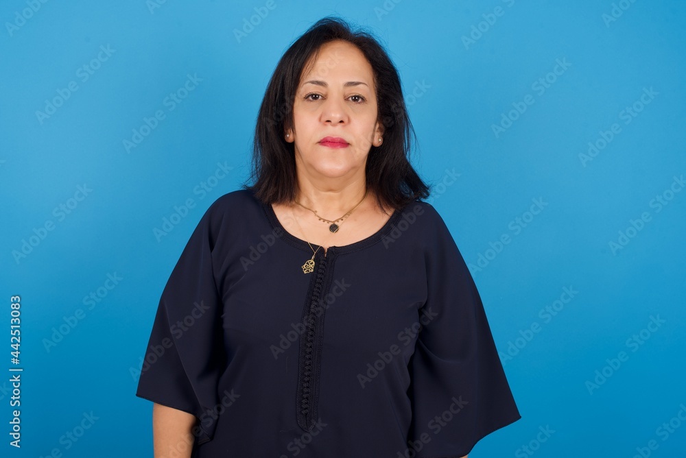 Fototapeta premium Joyful middle aged Arab woman standing against blue background looking to the camera, thinking about something. Both arms down, neutral facial expression.