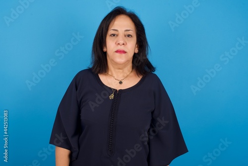 Joyful middle aged Arab woman standing against blue background looking to the camera, thinking about something. Both arms down, neutral facial expression.