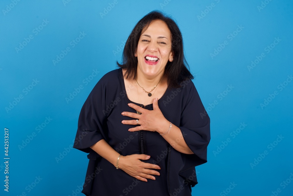middle aged Arab woman standing against blue background keeps hands crossed, laughs out loud at good joke, wears casual clothes. middle aged Arab woman standing against blue background keeps hands crossed, laughs out loud at good joke, wears casual clothes.