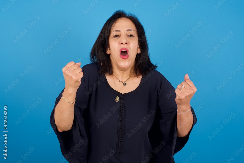 Joyful excited lucky middle aged Arab woman standing against blue background cheering, celebrating success, screaming yes with clenched fists Joyful excited lucky middle aged Arab woman standing against blue background cheering, celebrating success, screaming yes with clenched fists