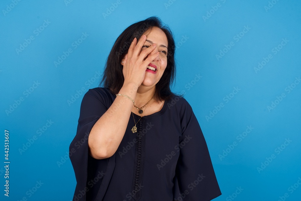 middle aged Arab woman standing against blue background makes face palm and smiles broadly, giggles positively hears funny joke poses middle aged Arab woman standing against blue background makes face palm and smiles broadly, giggles positively hears funny joke poses