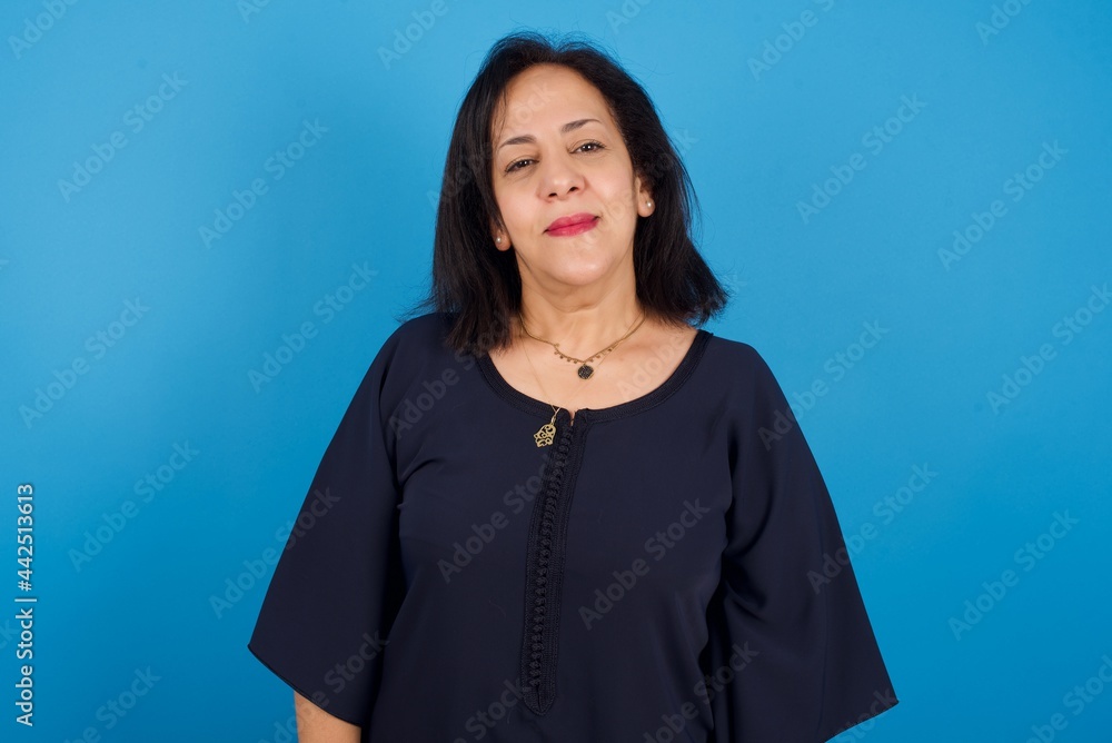 Fototapeta premium Happy middle aged Arab woman standing against blue background looking at camera with charming cute smile.