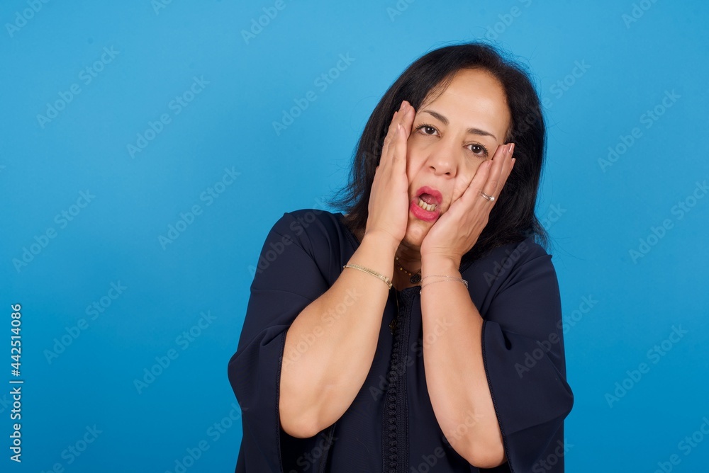middle aged Arab woman standing against blue background keeps hands on cheeks has bored displeased expression. Stressed hopeless model middle aged Arab woman standing against blue background keeps hands on cheeks has bored displeased expression. Stressed hopeless model