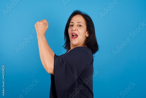 Overjoyed middle aged Arab woman standing against blue background glad to receive good news, clenching fist and making winning gesture. photo