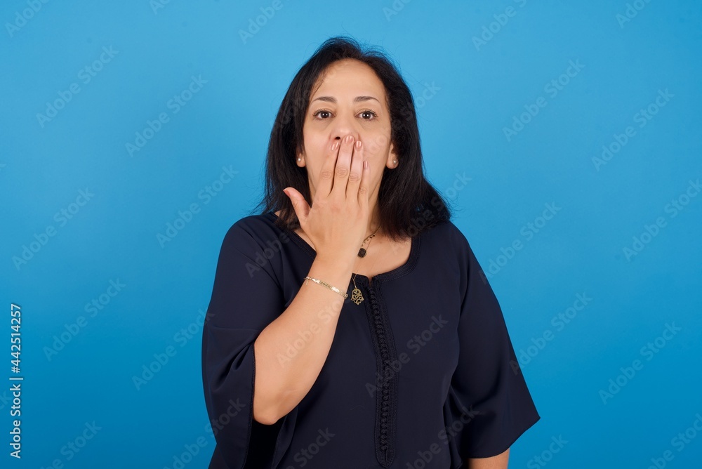 Oh! I think I said it! Close up portrait middle aged Arab woman standing against blue background cover open mouth by hand palm, look at camera with big eyes. Oh! I think I said it! Close up portrait middle aged Arab woman standing against blue background cover open mouth by hand palm, look at camera with big eyes.