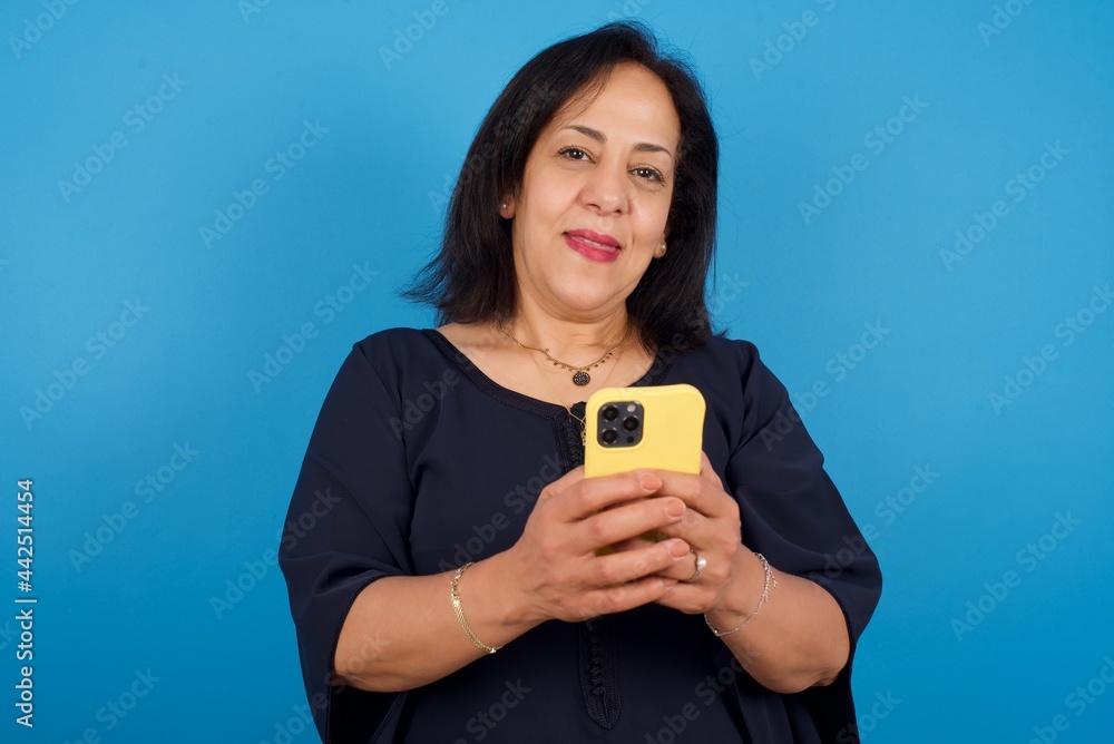 middle aged Arab woman standing against blue background Mock up copy space. Using mobile phone, typing sms message middle aged Arab woman standing against blue background Mock up copy space. Using mobile phone, typing sms message