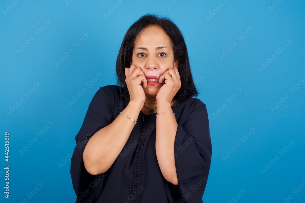 Terrified middle aged Arab woman standing against blue background looks empty space home alone moonless night Terrified middle aged Arab woman standing against blue background looks empty space home alone moonless night