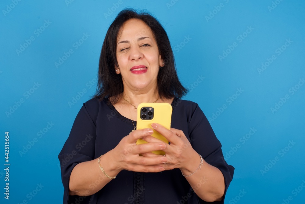 Pleased middle aged Arab woman standing against blue background using self phone and looking and winking at the camera. Flirt and coquettish concept. Pleased middle aged Arab woman standing against blue background using self phone and looking and winking at the camera. Flirt and coquettish concept.