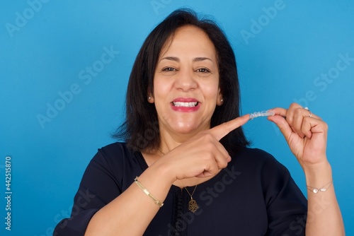 Fototapeta Naklejka Na Ścianę i Meble -  middle aged Arab woman standing against blue background holding an invisible aligner and pointing at it. Dental healthcare and confidence concept.
