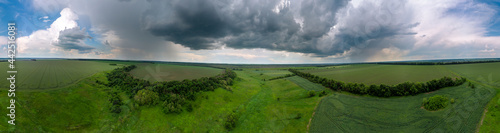 Aerial wide panorama summer view, fly above green fields with stormy cloudscape. Dark rainy scenic heavy weather in rural agriculture area. Drone view