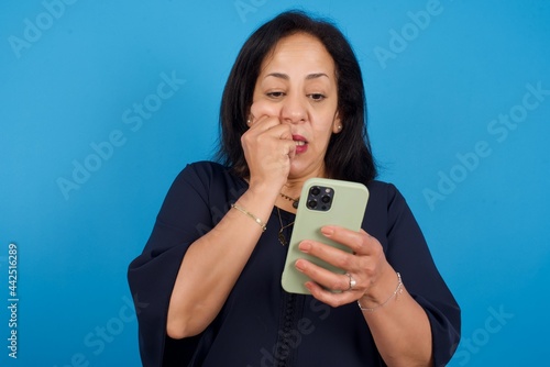 Portrait of pretty frightened middle aged Arab woman standing against blue background chatting biting nails after reading some scary news on her smartphone.