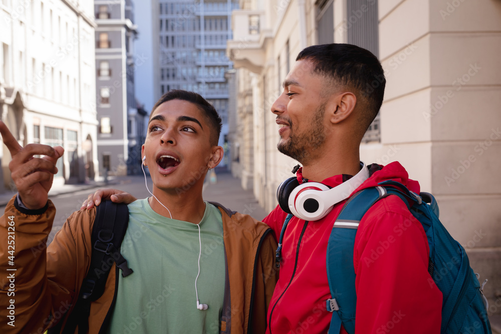 Two happy mixed race male friends with backpacks standing in city street talking Two happy mixed race male friends with backpacks standing in city street talking