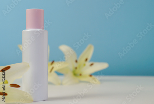 White blank cosmetic bottle with cream with pink cap, moisturizing lotion or shampoo framed with fresh lily flowers on a light blue background. Natural organic spa. Cosmetic concept. Front view.