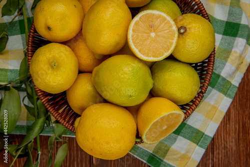 top view of fresh ripe lemons in a wicker basket with green leaves on plaid fabric background