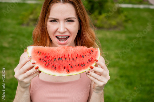 Close-up portrait of red-haired young woman with braces eating watermelon outdoors