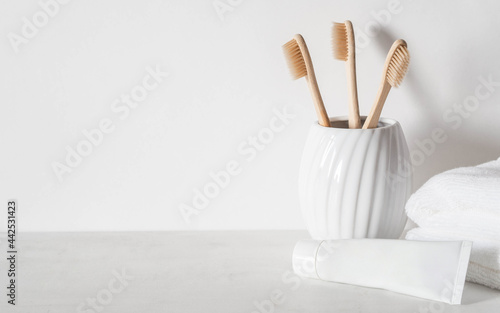 Minimal bath background with bamboo toothbrushs in ceramic glass, white tube of toothpaste and white towels.