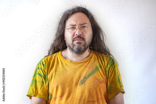 The man looks forward, squinting intently. A casually and sloppy dressed man with a beard and long hair in a paint-stained T-shirt on a light background.