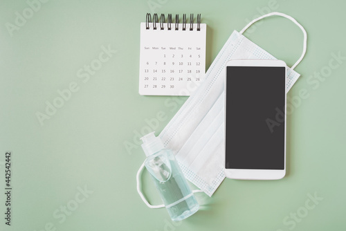 smart phone with clipping path on touchscreen on face mask, bottle of alcohol gel and opened calendar with green background ,new normal lifestyle, home isolation concept